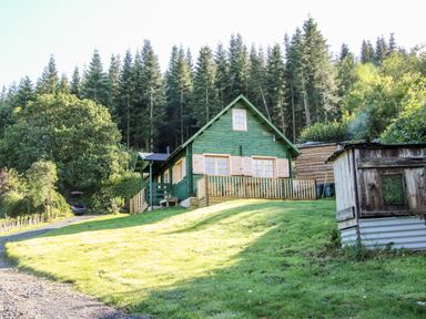 A green wooden cabin with a fenced porch on a grassy slope surrounded by trees and a gravel driveway with a parked car at Callow Fold in Dinchope near Craven Arms