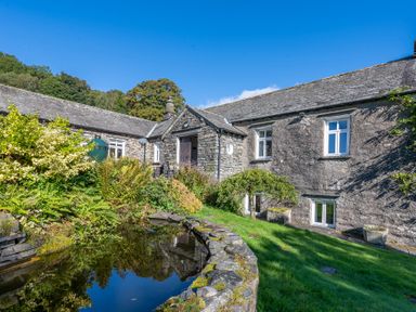 A stone house with white framed windows next to a garden pond and greenery at Field Head House in Graythwaite near Hawkshead