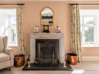 A fireplace with a mirror clock and dog statues between two windows with floral curtains at Field Head House Graythwaite near Hawkshead