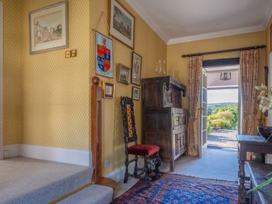 A hallway with patterned yellow wallpaper a dark wooden chair a wooden cabinet paintings on the wall and an open door showing an outdoor view at Field Head House in Graythwaite near Hawkshead