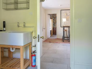 A room with a white sink on a wooden stand and fire extinguisher next to a doorway leading to another room with a rug and tall stool at Field Head House Graythwaite near Hawkshead
