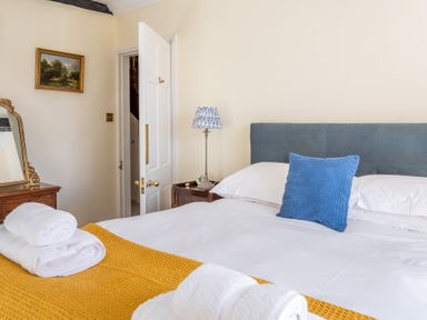 A bedroom with a bed covered in white sheets and a yellow blanket with folded white towels a blue cushion on the bed a small wooden bedside table with a lamp and a painting on the wall at Field Head House in Graythwaite near Hawkshead