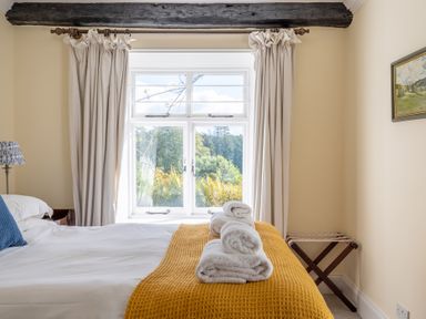 A bedroom with a bed covered with white sheets and a yellow blanket with folded towels on top next to a window with curtains and a wooden beam at Field Head House in Graythwaite near Hawkshead