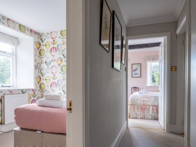 A hallway with framed pictures on the wall leading to two bedrooms one with hot air balloon wallpaper and pink bedspread the other with bird artwork and floral bedspread at Field Head House in Graythwaite near Hawkshead