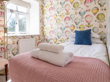 A bedroom with a bed covered in white and pink bedding with a blue pillow and towels on top and hot air balloon wallpaper at Field Head House in Graythwaite near Hawkshead
