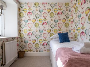 A bedroom with a single bed with white bedding a blue pillow and pink blanket hot air balloon wallpaper and a window with a radiator underneath at Field Head House in Graythwaite near Hawkshead