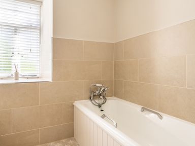 A bathroom with a white bathtub beige tiled walls and a window with white blinds at Field Head House Graythwaite near Hawkshead