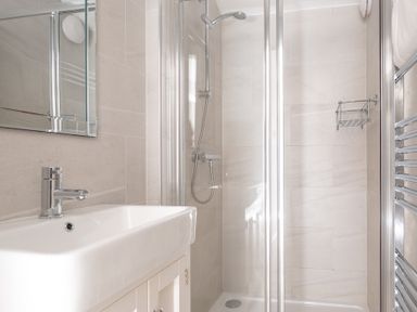 A bathroom with a white sink and faucet a mirrored cabinet a glass-enclosed shower with a handheld showerhead a chrome towel rack and beige tiled walls at Field Head House in Graythwaite near Hawkshead