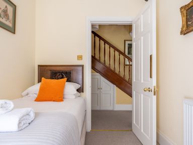 A bedroom with a single bed with an orange pillow and folded towels on the bed a white door open showing a wooden staircase and storage cabinets at Field Head House in Graythwaite near Hawkshead