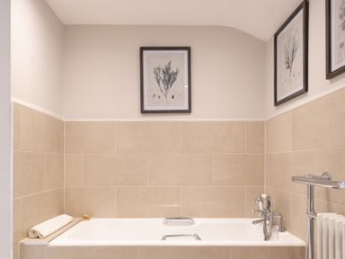 A bathroom with a white bathtub beige tiled walls and framed botanical prints above the tub at Field Head House in Graythwaite near Hawkshead
