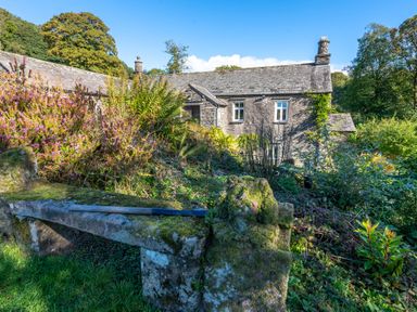 An old stone house surrounded by dense greenery and flowers with a moss covered stone bench and a gardening tool in front at Field Head House in Graythwaite near Hawkshead