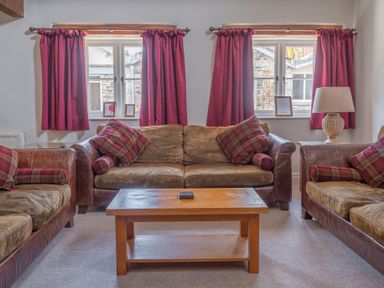 A living room with three brown sofas with plaid cushions red curtains on two windows and a wooden coffee table at Grubbings Graythwaite near Hawkshead