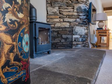 A room with a stone fireplace wood burning stove and a table with lamp at Grubbings Graythwaite near Hawkshead