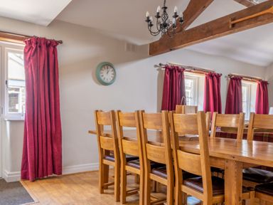 A dining room with a long wooden table and chairs under a wooden beam and chandelier with red curtains at Grubbings Graythwaite near Hawkshead