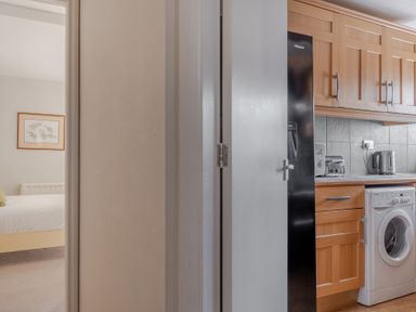 A kitchen with wooden cabinets washing machine and black refrigerator adjacent to a bedroom with a bed and framed artwork at Grubbings Graythwaite near Hawkshead