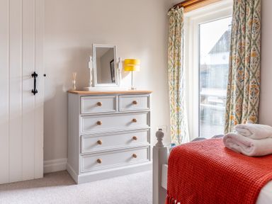 A bedroom with a white dresser with mirror and yellow lamp next to a window with floral curtains and a bed with a red blanket stacked towels at Grubbings Graythwaite near Hawkshead