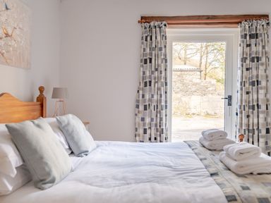 A bedroom with a wooden bed with pillows and towels near a glass door with patterned curtains at Grubbings in Graythwaite near Hawkshead