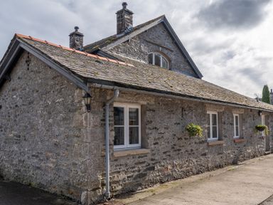 A stone house with a mossy tiled roof and white windows with hanging flower baskets at Grubbings in Graythwaite near Hawkshead