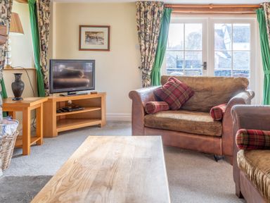 A living room with a wooden coffee table sofa with plaid pillows television on a wooden stand and large windows with floral and green curtains at Bull Pen Graythwaite near Hawkshead