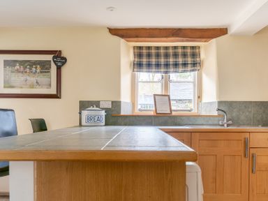 A kitchen counter with a bread container near a window with plaid curtain and wooden cabinets at Bull Pen in Graythwaite near Hawkshead