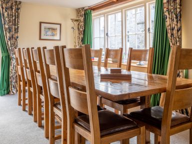 A dining room with a long wooden table and chairs near windows with green and patterned curtains at Bull Pen in Graythwaite near Hawkshead