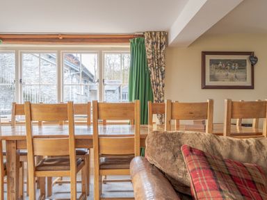A dining area with a wooden table and chairs next to large windows and a sofa with a plaid cushion at Bull Pen in Graythwaite near Hawkshead