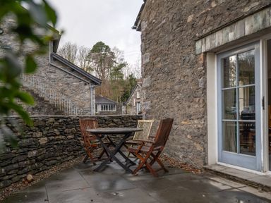 A stone patio with a round table and three wooden chairs outside a building with stone walls and a glass door at Bull Pen in Graythwaite near Hawkshead