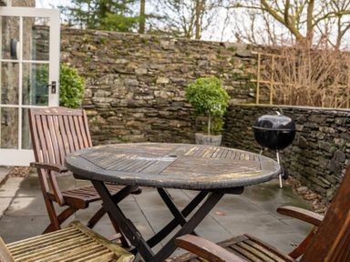 A patio with wooden chairs and a round wooden table near a stone wall with a charcoal grill and potted plants at Bull Pen in Graythwaite near Hawkshead