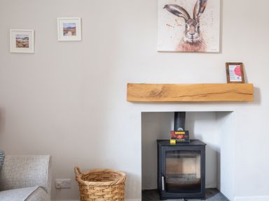 A fireplace with a black stove and a wooden mantle with a rabbit painting above and a wicker basket next to a gray sofa at Briar in Graythwaite near Hawkshead
