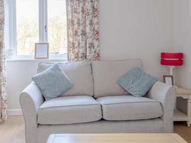 A seating area with a light grey sofa with two blue patterned cushions a side table with a red lamp and a window with floral curtains at Briar Graythwaite near Hawkshead