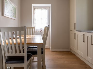 A dining area with a wooden table and four chairs with cushions next to a door with a sheer curtain and kitchen cabinets at Briar Graythwaite near Hawkshead