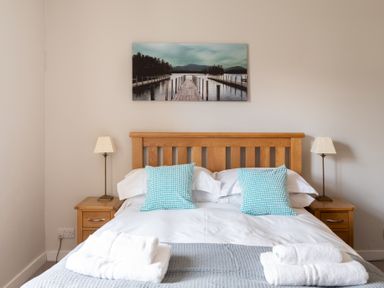 A bedroom with a wooden double bed white bedding blue checkered cushions bedside tables with lamps and a landscape picture above the bed at Briar Graythwaite near Hawkshead