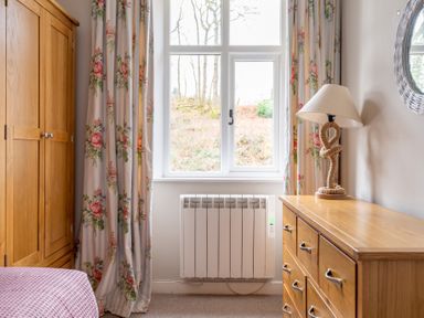 A bedroom corner with a wooden wardrobe a chest of drawers with a lamp floral curtains and a window at Briar Graythwaite near Hawkshead
