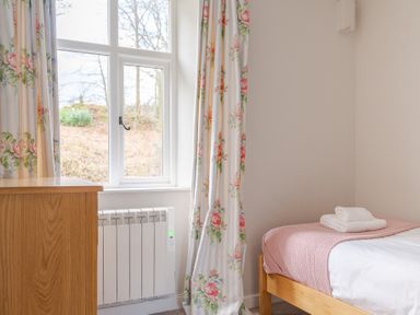 A bedroom with a window with floral curtains a wooden dresser a bed with folded towels and a pink blanket at Briar Graythwaite near Hawkshead