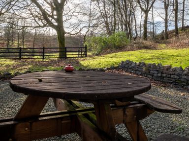 A round wooden picnic table with a red container on top in a garden with gravel ground grass trees and a stone wall at Briar Graythwaite near Hawkshead