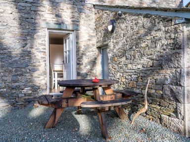 A stone patio with a wooden round picnic table and attached benches outside a building with an open door showing a dining room at Briar Graythwaite near Hawkshead