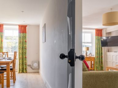 A dining area with wooden table and chairs and a living room with sofa and TV visible through an open door at Eel House Cottage in Graythwaite Estate near Hawkshead