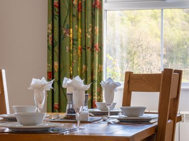 A dining table set with white plates bowls and glasses with napkins near a window with floral curtains at Eel House Cottage in Graythwaite Estate near Hawkshead