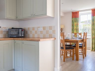 A kitchen corner with cabinets and a microwave next to a dining area with a wooden table and chairs with green and orange curtains at Eel House Cottage Graythwaite Estate near Hawkshead