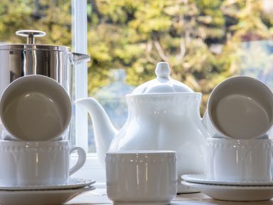 A white teapot with stacked white cups and saucers on a windowsill with a blurred outdoor view at Eel House Cottage Graythwaite Estate near Hawkshead