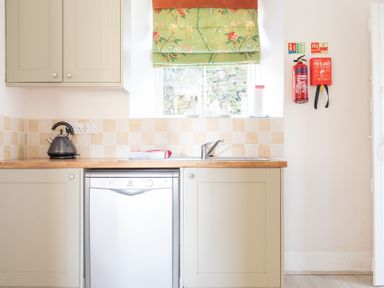 A kitchen with a kettle on the counter a dishwasher a sink and a window with floral curtain at Eel House Cottage Graythwaite Estate near Hawkshead