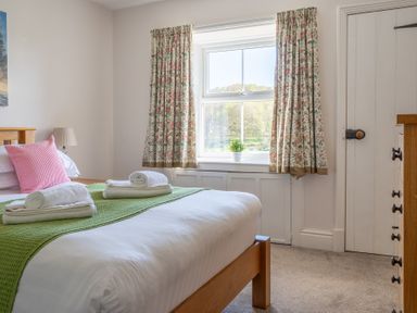 A bedroom with a wooden bed a pink pillow folded towels a window with floral curtains and a wooden dresser at Eel House Cottage Graythwaite Estate near Hawkshead