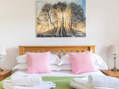 A bedroom with a wooden bed frame pink pillows white towels bedside lamps and a painting of trees at Eel House Cottage Graythwaite Estate near Hawkshead