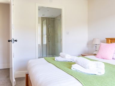 A bedroom with a bed covered with a green throw and white towels with a pink pillow and an open door to a bathroom with a glass shower at Eel House Cottage in Graythwaite Estate near Hawkshead