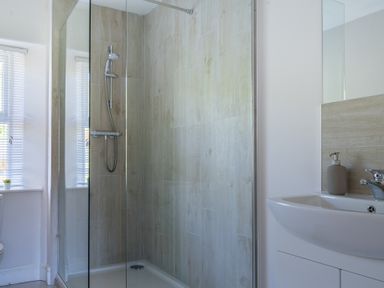 A bathroom with a glass shower enclosure a showerhead a white sink with a soap dispenser and windows with blinds at Eel House Cottage in Graythwaite Estate near Hawkshead