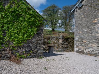 Outdoor area between stone buildings with gravel ground a wooden chair and stone wall at Eel House Cottage in Graythwaite Estate near Hawkshead