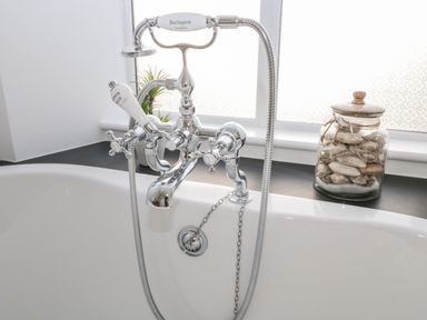 A bathtub with a chrome faucet and handheld shower head near a window with a jar of shells and a small plant on the windowsill at Pottergill in Windermere
