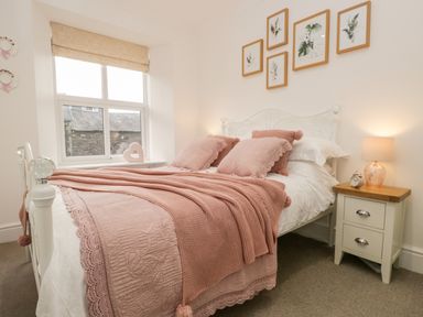 A bedroom with a white metal bed frame pink bedding a wooden bedside table with a lamp and alarm clock and framed botanical prints on the wall at Pottergill in Windermere