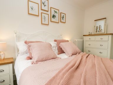 A bedroom with a white metal bed frame pink pillows and blankets a white nightstand with a lamp and a white dresser with a mirror on top at Pottergill in Windermere