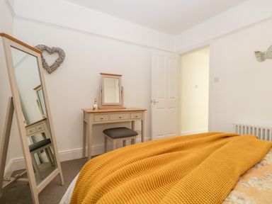 A bedroom with a bed covered by a mustard yellow blanket a dressing table with a mirror and stool a full-length mirror on stand a heart shaped wall decoration and an open door at Pottergill in Windermere